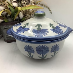 May include: A round ceramic casserole dish with a matching lid, featuring a white base with hand-painted blue cornflower motifs. The bowl has two side handles and a blue rim, while the lid includes a green knob and a blue-painted edge.