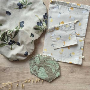 Peut inclure: Ensemble d'articles de cuisine réutilisables, comprenant des couvercles de bols et des sacs à provisions, avec des motifs d'olives et de poires. Les articles sont en tissu beige avec des branches d'olives vertes et noires et des poires jaunes. Sont également inclus des dessous de verre hexagonaux verts et marron.