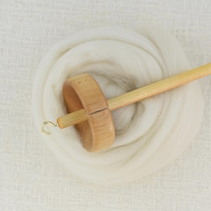 K&ouml;nnte beinhalten: Eine Handspindel aus Holz mit einem hellen Schaft und einem runden Wirtel, die an der Spitze mit einem kleinen Metallhaken versehen ist. Die Spindel liegt auf einem gewickelten B&uuml;ndel aus weicher, wei&szlig;er, ungesponnener Wolle und zeigt Werkzeug.