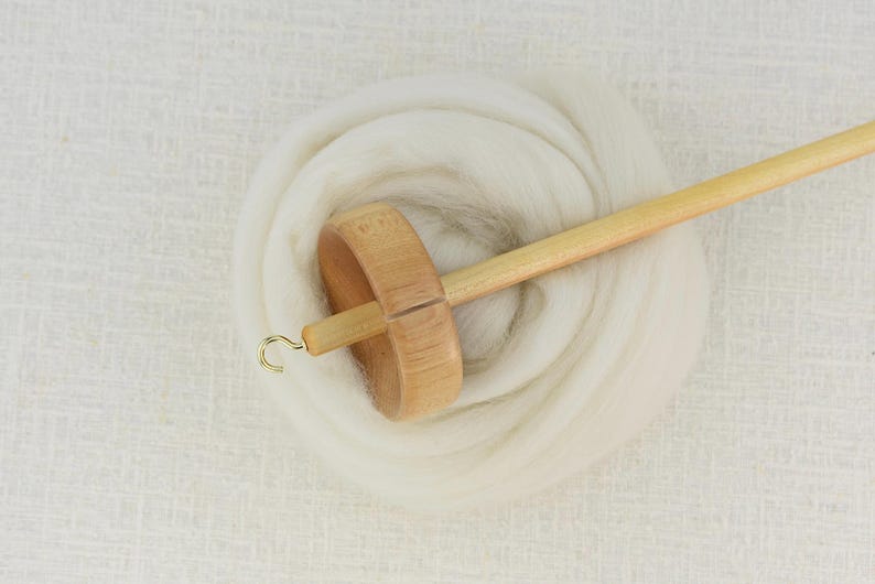 K&ouml;nnte beinhalten: Eine Handspindel aus Holz mit einem hellen Schaft und einem runden Wirtel, die an der Spitze mit einem kleinen Metallhaken versehen ist. Die Spindel liegt auf einem gewickelten B&uuml;ndel aus weicher, wei&szlig;er, ungesponnener Wolle und zeigt Werkzeug.
