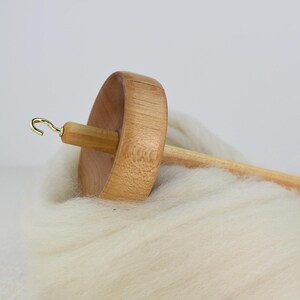 K&ouml;nnte beinhalten: Eine Holzspindel mit einem hellbraunen, kreisf&ouml;rmigen Wirtel und einem langen Schaft, der mit einem kleinen goldfarbenen Metallhaken versehen ist. Die Spindel liegt auf einem weichen, dicken B&uuml;ndel aus ungesponnener, cremewei&szlig;er Wolle.