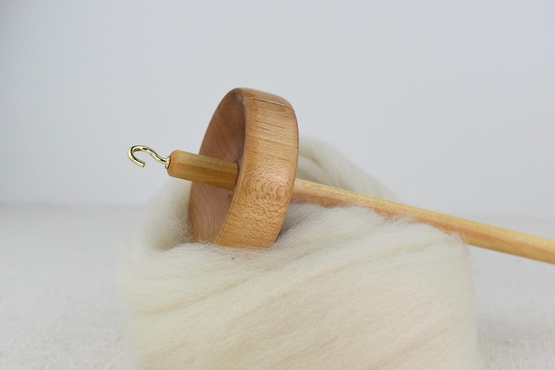 K&ouml;nnte beinhalten: Eine Holzspindel mit einem hellbraunen, kreisf&ouml;rmigen Wirtel und einem langen Schaft, der mit einem kleinen goldfarbenen Metallhaken versehen ist. Die Spindel liegt auf einem weichen, dicken B&uuml;ndel aus ungesponnener, cremewei&szlig;er Wolle.