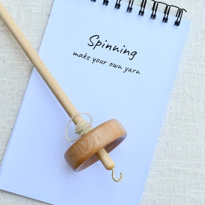 K&ouml;nnte beinhalten: Eine Holzspindel mit einem hellen Wirtel und einem Metallhaken an der Spitze, platziert auf einem wei&szlig;en Spiralblock. Der Notizblock enth&auml;lt den aufgedruckten Text "Spinning" und "make your own yarn" in einer einfachen schwarzen Schrift.