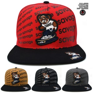 May include: A red, black, and brown baseball cap with a cartoon bear wearing a white shirt with the word "SAVAGE" printed on it. The bear is skateboarding and the cap is covered in the word "SAVAGE" repeated in black.