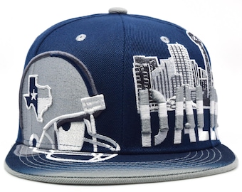 Op de afbeelding: Een marineblauwe baseballcap met een grijze klep en een grijze bovenknop. De cap heeft een geborduurd footballhelm met de omtrek van de staat Texas en een ster, en het woord "DALLAS" in grijs, samen met een stadsgezicht.