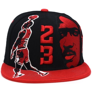 Pode incluir: Um boné de beisebol preto com aba reta vermelha e um botão vermelho no topo. A frente apresenta uma silhueta vermelha bordada de um jogador de basquete em movimento, o grande número 23 vermelho e um retrato vermelho detalhado de um rosto.