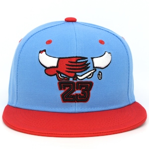 May include: A light blue baseball cap with a red brim and a red button on top. The front features an embroidered red and white bull logo with the number 23 in black and red outline centered directly below the bull graphic on the crown.