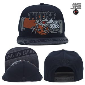 Puede incluir: Una gorra snapback negra con un bordado que muestra una mano marrón estrechando una cabeza de serpiente blanca. El texto "TRUST NO ONE" está bordado en gris en la parte delantera e impreso en la visera. Incluye una correa de plástico ajustable.