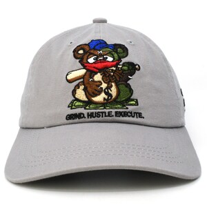 May include: A gray baseball cap featuring an embroidered cartoon bear holding a money bag. The bear has a blue hat, red mask, and green and brown fur. The text "GRIND. HUSTLE. EXECUTE." is embroidered below the bear.