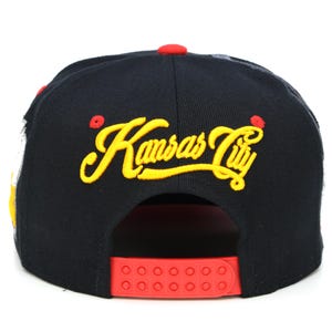 May include: Black baseball cap with a red and yellow embroidered "Kansas City" logo. The cap has a red adjustable strap with small holes.