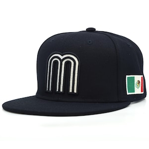 May include: A dark navy blue baseball cap featuring a large, white, embroidered letter M on the front panel. The side of the cap displays a rectangular patch of the Mexican flag, which includes green, white, and red vertical stripes with an emblem.