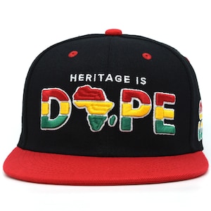 May include: Black snapback hat with a red brim. The hat features the embroidered text "HERITAGE IS DOPE" with a colorful map of Africa. The letters are in red, yellow, green, and white.