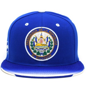 Könnte beinhalten: Eine blaue und weiße Baseballkappe mit dem Staatswappen von El Salvador, das auf der Vorderseite gestickt ist. Das Wappen zeigt einen Schild mit einem Vulkan, einer Sonne und einem See. Der Text "Presidencia de la Republica El Salvador" ist um den Schild herum gestickt.