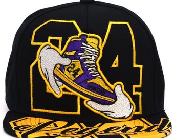 Pode incluir: Boné de beisebol preto com um gráfico de tênis amarelo e roxo segurado por mãos enluvadas brancas. O número 24 está presente na lateral. A aba tem a palavra "Legend" em amarelo.