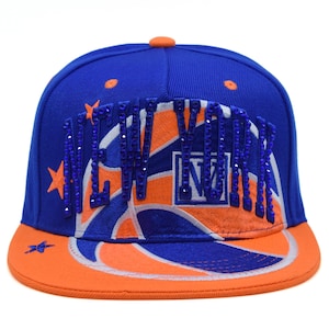 May include: A blue and orange baseball cap featuring the text NEW YORK in blue rhinestones across the front. The cap displays an embroidered basketball graphic with orange and blue panels, accented by small orange and blue star shapes.