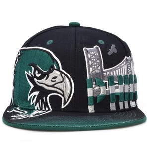 Può includere: Un cappellino da baseball nero con una testa d'aquila verde ricamata sul lato sinistro e un'illustrazione di un ponte grigio con il testo PHIL sul lato destro. Il cappellino presenta una visiera verde scuro e occhielli verdi sulla corona.