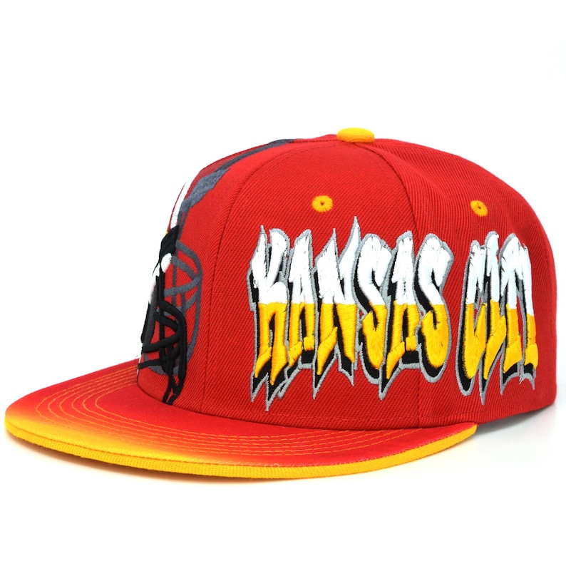 May include: A red and yellow baseball cap with a white and yellow "Kansas City" graphic on the front.