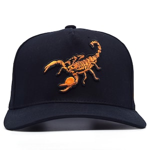 May include: A black baseball cap featuring an embroidered orange scorpion design. The scorpion is detailed with a textured appearance, and the cap has a curved brim. The cap is a classic, adjustable style.