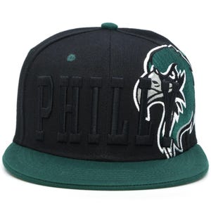 Könnte beinhalten: Schwarze und grüne Baseballkappe mit einem großen gestickten "PHILL"-Text und einer grünen Vogelgrafik an der Seite.