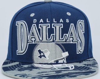 Op de afbeelding: Een marineblauwe baseballcap met het woord "DALLAS" in grote, zilverkleurige geborduurde letters. De cap heeft een grijze klep met een patroon en een logo van een footballhelm met de omtrek van de staat Texas.