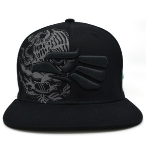 Puede incluir: Gorra de b&eacute;isbol negra con un dise&ntilde;o bordado en gris de un &aacute;guila y una mano estilizada en la parte delantera. El dise&ntilde;o est&aacute; en el lado izquierdo de la gorra, y el lado derecho es de color negro s&oacute;lido.