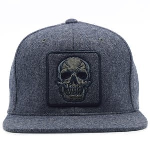 May include: A charcoal grey baseball cap with a flat brim and a square patch on the front panel. The patch features an embroidered olive green skull design with black detailing, centered on the crown of the cap. The fabric has a felt-like texture.