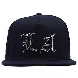 Może przedstawiać: Czarna czapka bejsbolowa z literami "LA" w gotyckiej czcionce. Litery są ozdobione małymi, błyszczącymi dżetami. Czapka ma płaski daszek i zaokrągloną koronę.