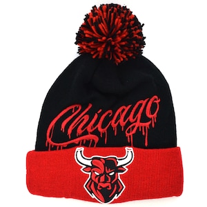 Könnte beinhalten: Schwarze und rote Strickmütze mit Bommel oben. Die Mütze hat ein rotes gesticktes Chicago-Logo mit tropfender Farbe und ein rotes gesticktes Stierkopf-Logo am Bündchen.