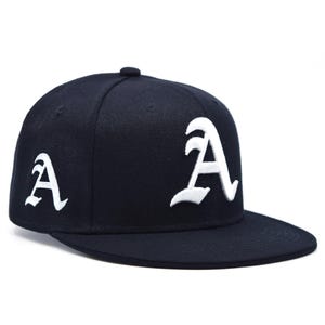 May include: A navy blue baseball cap with a flat peak. The cap has a white embroidered letter "A" on the front and side. The cap has a button on top.