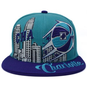 May include: A teal blue and purple baseball cap with a city skyline and basketball graphic. The cap has the word "CHA" and "Charlotte" embroidered on it.