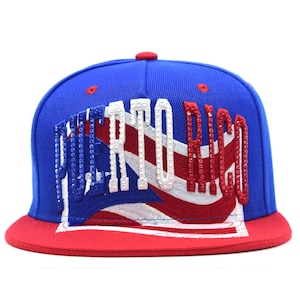 May include: A blue baseball cap with a red brim featuring an embroidered Puerto Rico flag design on the front. The words PUERTO RICO are displayed in large, raised, rhinestone-encrusted letters, with blue, white, and red stones matching the flag.