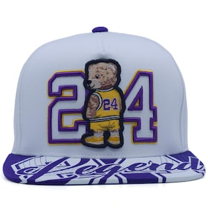 May include: A light grey baseball cap featuring a large embroidered number 24 in purple and yellow. Centered is an embroidered teddy bear wearing a yellow basketball jersey with the number 24. The purple brim displays the word Legend in white.