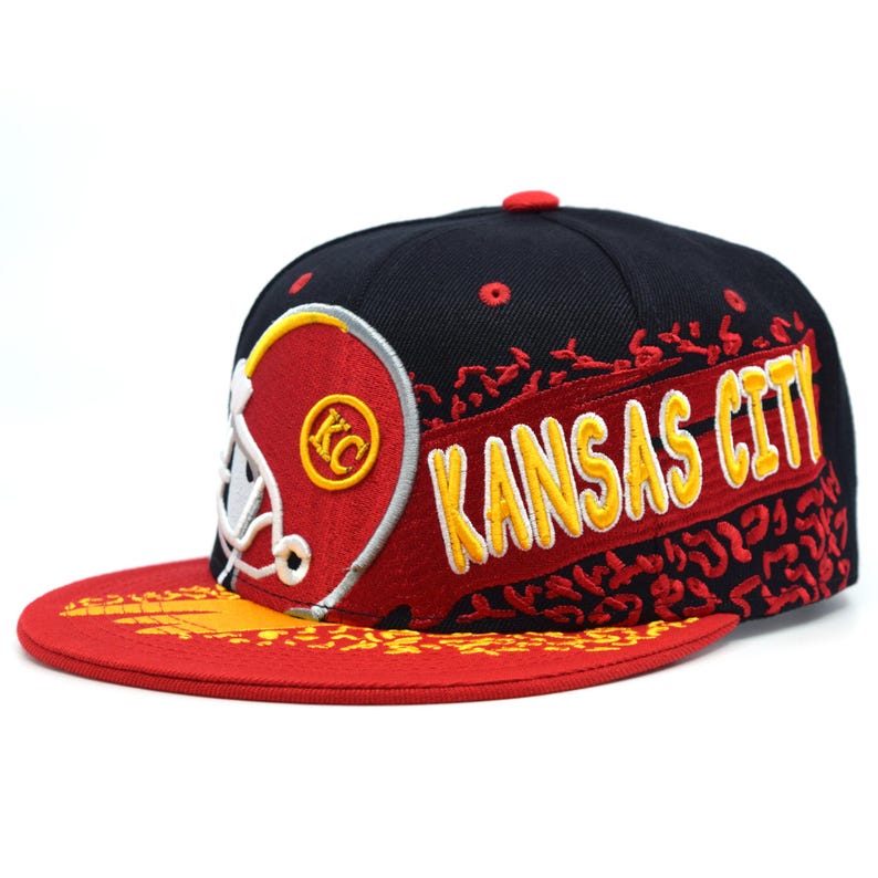 以下が含まれることがあります： 黄色い文字で「KANSAS CITY」と書かれた黒と赤の野球帽。赤いツバと赤と黄色のフットボールヘルメットのデザインが特徴です。つばは平らで、上部に赤いボタンが付いています。