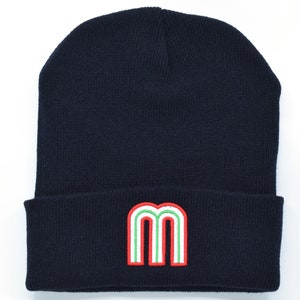 Könnte beinhalten: Eine marineblaue Strickmütze mit einem aufgestickten roten, grünen und weißen Buchstaben "M" auf der Vorderseite.