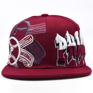 May include: A maroon baseball cap with white and grey embroidered details. The cap features the word "PHILLIES" in white and maroon, along with baseball-themed patches and the word "BASEBALL".