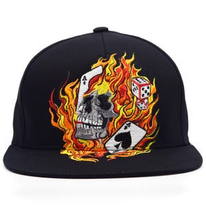 Peut inclure: Une casquette de baseball noire ornée d'une broderie représentant un crâne gris entouré de flammes orange et jaunes vives, accompagnée de deux cartes à jouer montrant l'as de pique et une paire de dés blancs à points noirs.