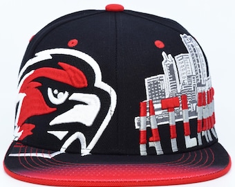 Könnte beinhalten: Schwarze Baseballkappe mit einem rot-weißen Vogellogo und einem Stadtlandschaftsdesign. Der Schirm ist rot mit einem Farbverlauf. Die Kappe hat einen flachen Schirm und einen roten Knopf oben. Durchmesser ca. 20 cm.