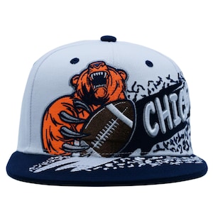 Puede incluir: Una gorra de béisbol blanca con visera y botón superior azul marino. El frente presenta un oso naranja bordado sosteniendo un balón de fútbol americano marrón, con el texto CHICAGO en letras blancas sobre un fondo diagonal azul marino.
