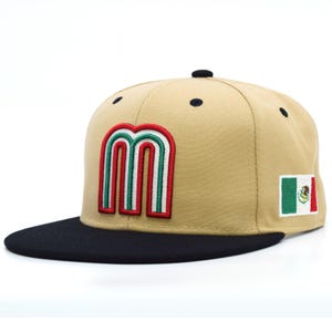 Op de afbeelding: Een beige honkbalpet met een platte zwarte klep en een zwarte knoop aan de bovenkant. De voorkant is voorzien van een grote, geborduurde letter M in rood, wit en groen. Een kleine rechthoekige patch van de Mexicaanse vlag is aan de zijkant genaaid.