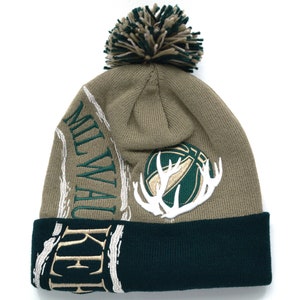 Puede incluir: Un gorro de punto verde y marrón con un pompón en la parte superior. El gorro tiene el logotipo de los Milwaukee Bucks en la parte delantera y la palabra "Milwaukee" en el lateral.