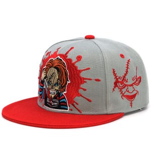 May include: A gray baseball cap with a red bill and top button. The cap features an embroidered image of a horror movie character with a knife, set against a red splatter design. The cap also has a stitched face design.