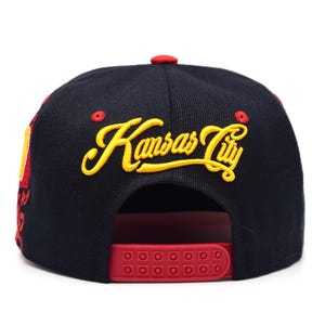 以下が含まれることがあります： 黒と赤の野球帽。黄色い筆記体で「Kansas City」と刺繍されています。赤いスナップバッククロージャーと赤いボタンが付いています。つばは黒で、赤いアクセントが付いています。