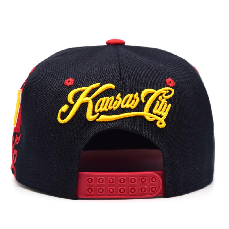 以下が含まれることがあります： 黒と赤の野球帽。黄色い筆記体で「Kansas City」と刺繍されています。赤いスナップバッククロージャーと赤いボタンが付いています。つばは黒で、赤いアクセントが付いています。