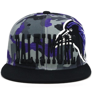 Może przedstawiać: Czarna i fioletowa czapka z daszkiem w kamuflażu z czarnym logo Baltimore Ravens i haftowanym napisem "Baltimore" w kolorze fioletowym z przodu.