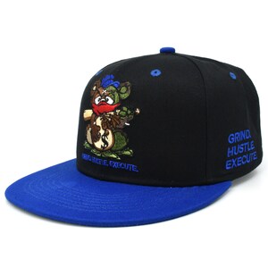 May include: A black and blue baseball cap with an embroidered cartoon bear holding a bag of money. The cap has the text "Grind Hustle Execute" on the front and "Grind Hustle Execute" on the back.