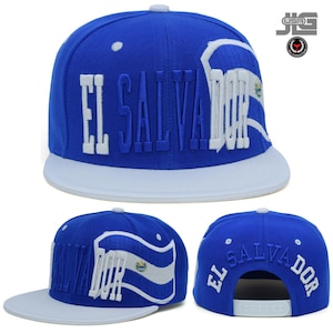 May include: A blue snapback cap featuring the text EL SALVADOR embroidered in white and blue. The design includes a stylised waving flag graphic with the national emblem. The cap has a white flat brim and an adjustable white plastic snap closure.
