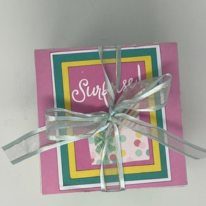 May include: A pink card with a green, yellow, and blue border. The card has a colorful design with the word "Surfrise!" written in cursive. The card is tied with a light blue ribbon with gold accents.