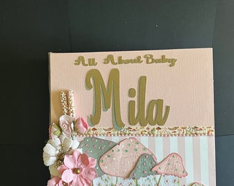 May include: A pink and white scrapbook titled "All About Baby" with the name "Mila" in gold lettering. The cover features paper flowers, mushrooms, and a picket fence design. The scrapbook is a baby-themed item.