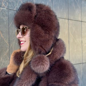 Puede incluir: Un sombrero de piel marrón con orejeras y un abrigo de piel a juego. El sombrero tiene forma redondeada y un detalle de pompón. El abrigo tiene un diseño en capas. La persona lleva gafas de sol y guantes.