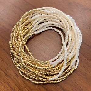 Puede incluir: Una cadena de cintura de cuentas doradas y blancas, también conocida como cuentas de cintura o cadena de vientre. La cadena está hecha de cuentas pequeñas y redondas y está diseñada para usarse alrededor de la cintura.